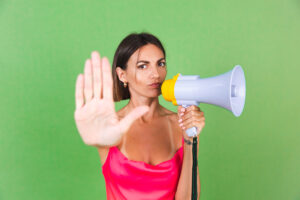 Stylish woman in pink silk dress on green background, with megaphone, isolated, show stop gesture with serious face